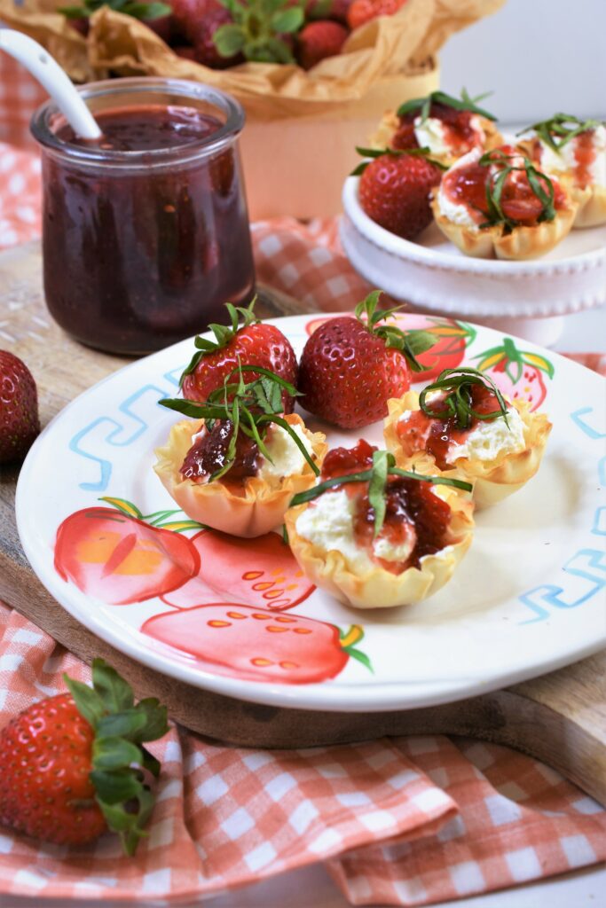 a strawberry printed ceramic plate with phyllo cups filled with cream cheese with red pepper strawberry jam drizzled on top of each one with chopped basil as garnish on top of cutting board