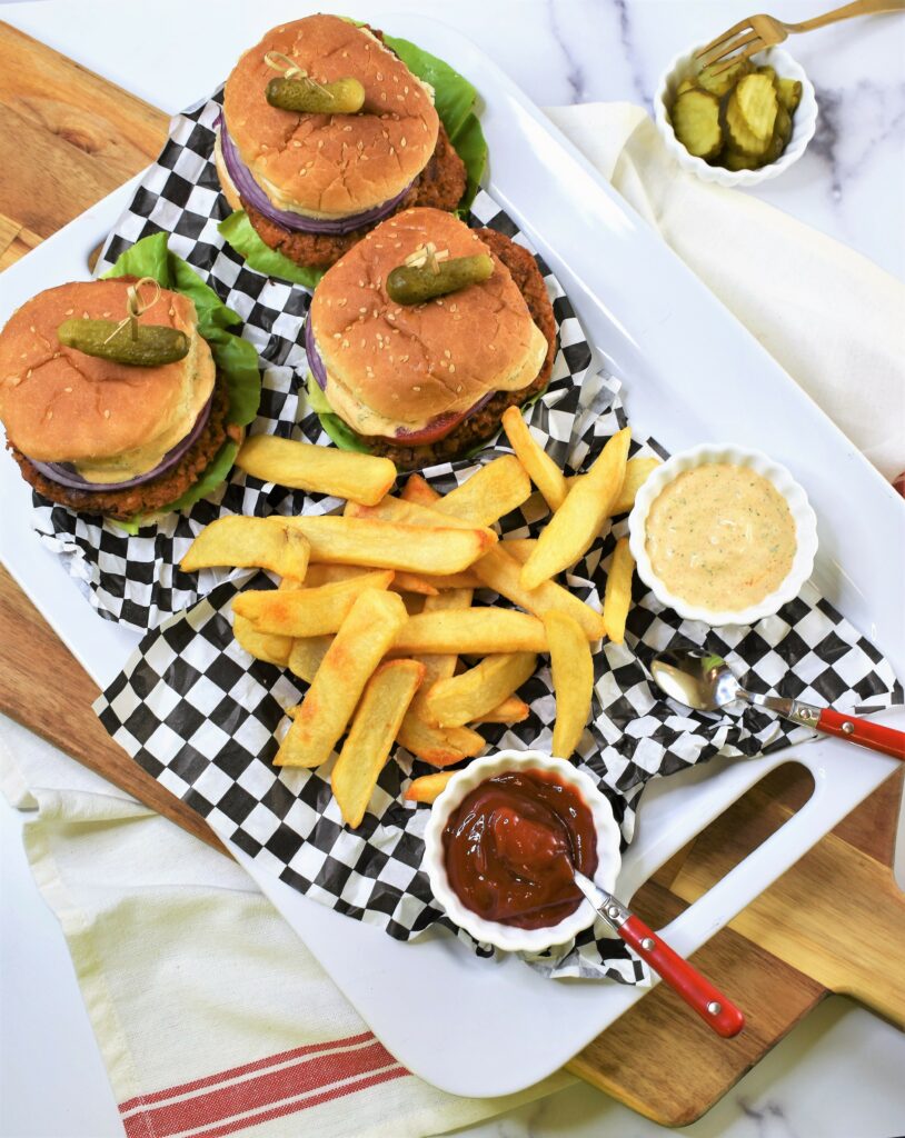 overhead image of large white rectangle ceramic platter of three Chipotle Butternut Squash Burger with Hatch Chile Aioli on black and white checkered deli paper with french fries and sides of ketchup and Hatch chile aioli with sliced bread and butter pickles in the background