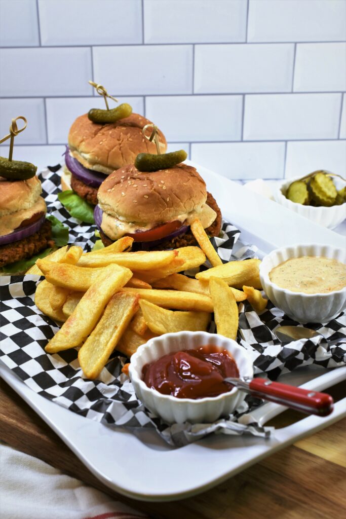 large white rectangle ceramic platter of three veggie burgers on black and white checkered deli paper with french fries and sides of ketchup and Hatch chile aioli with sliced bread and butter pickles in the background
