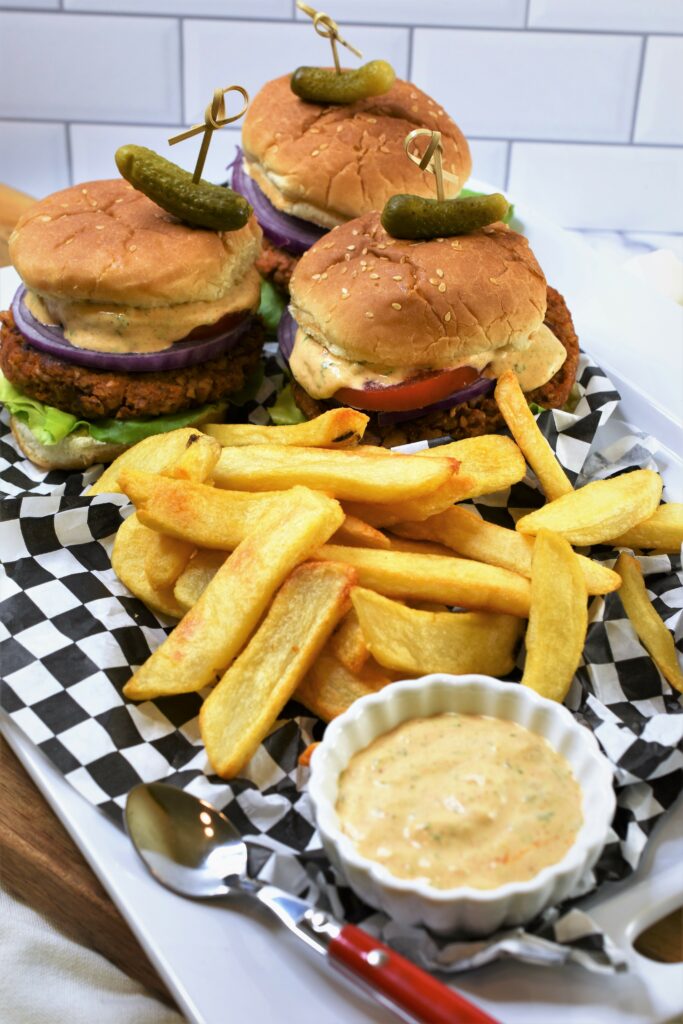 3 Chipotle Butternut Squash Burgers with Hatch Chile Aioli on a white ceramic platter on top of black and white checkered paper with thick french fries on cutting board with subway tile in the background