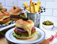 up close image of 1 Chipotle Butternut Squash Burger with others in the background with Hatch Chile Aioli on a blue and white plate with french fries in galvanized steel cup