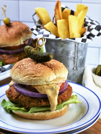 up close image of 1 Chipotle Butternut Squash Burger with others in the background with Hatch Chile Aioli on a blue and white plate with french fries in galvanized steel cup