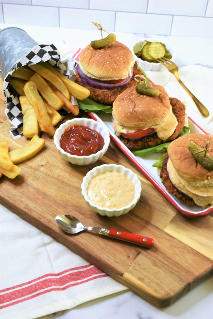 set of 3 Butternut Squash Burgers with Hatch Chile Aioli on white and red enamelware tray with sides of ketchup and Hatch chile aioli on cutting board with steak fries spilling out of galvanized cup on its side