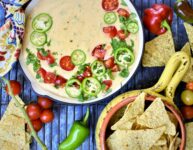 Hatch Chile Queso Blanco served in a red cast iron skillet with colorful napkin tied around handle with fresh cherry tomatoes and sliced jalopenos with tortilla chips
