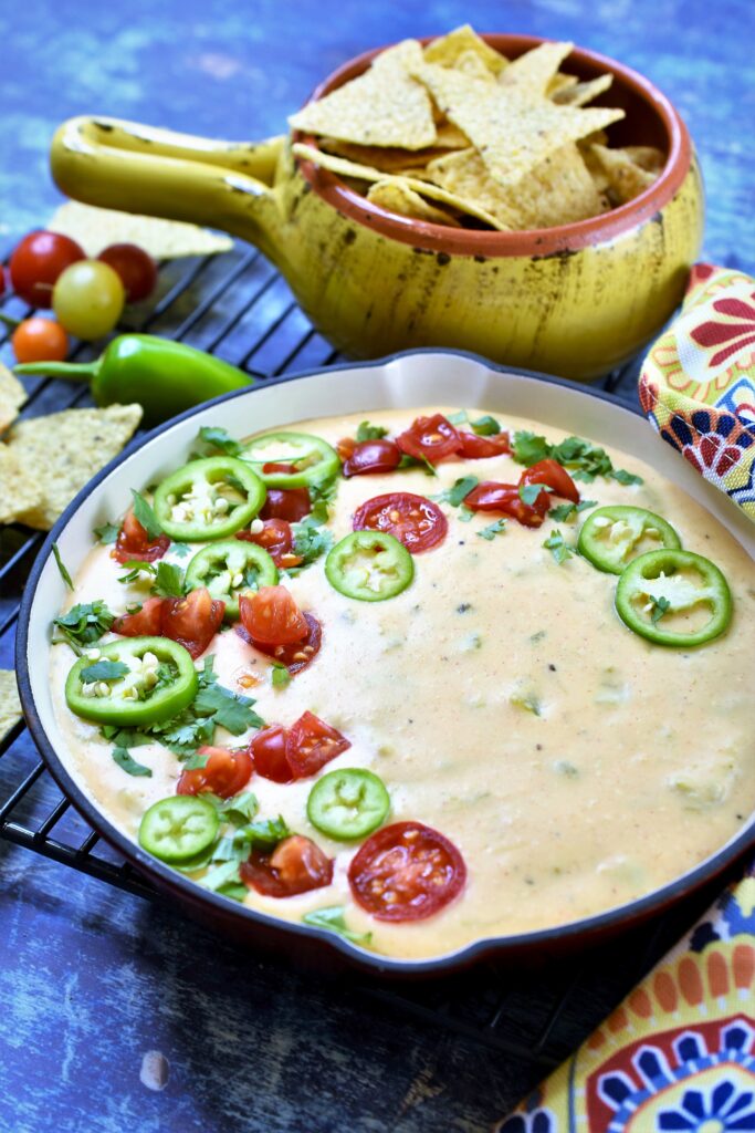 warm cheese dip served in a red cast iron skillet with colorful napkin tied around handle with fresh cherry tomatoes and sliced jalapenos with tortilla chips