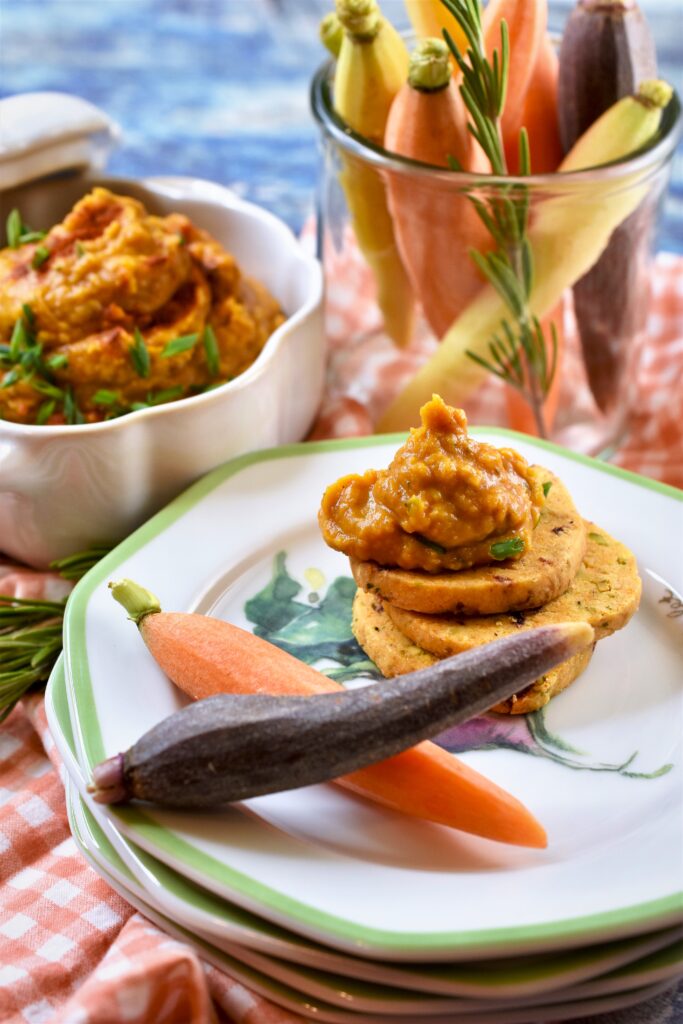 white and green appetizer plates with a stack of crackers with pumpkin bean dip on them alongside multi colored carrots